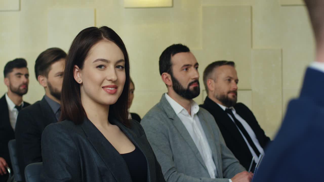 joven mujer de negocios caucásica sentada en una silla entre la gente en una sala de conferencias, luego gira la cara hacia la cámara y sonríe