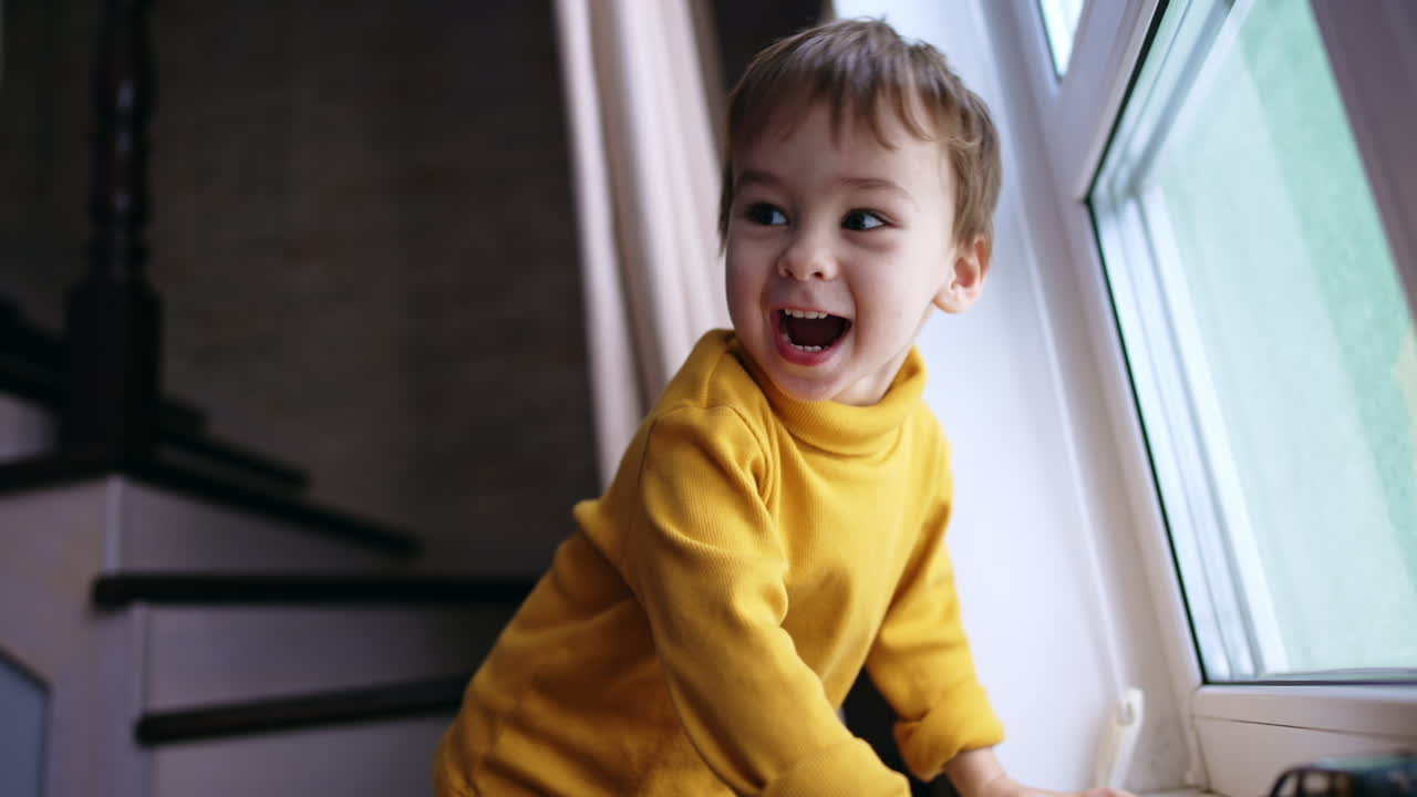 Beautiful toddler boy standing near the window. Smiling kid looking back and hopping. Low angle view.