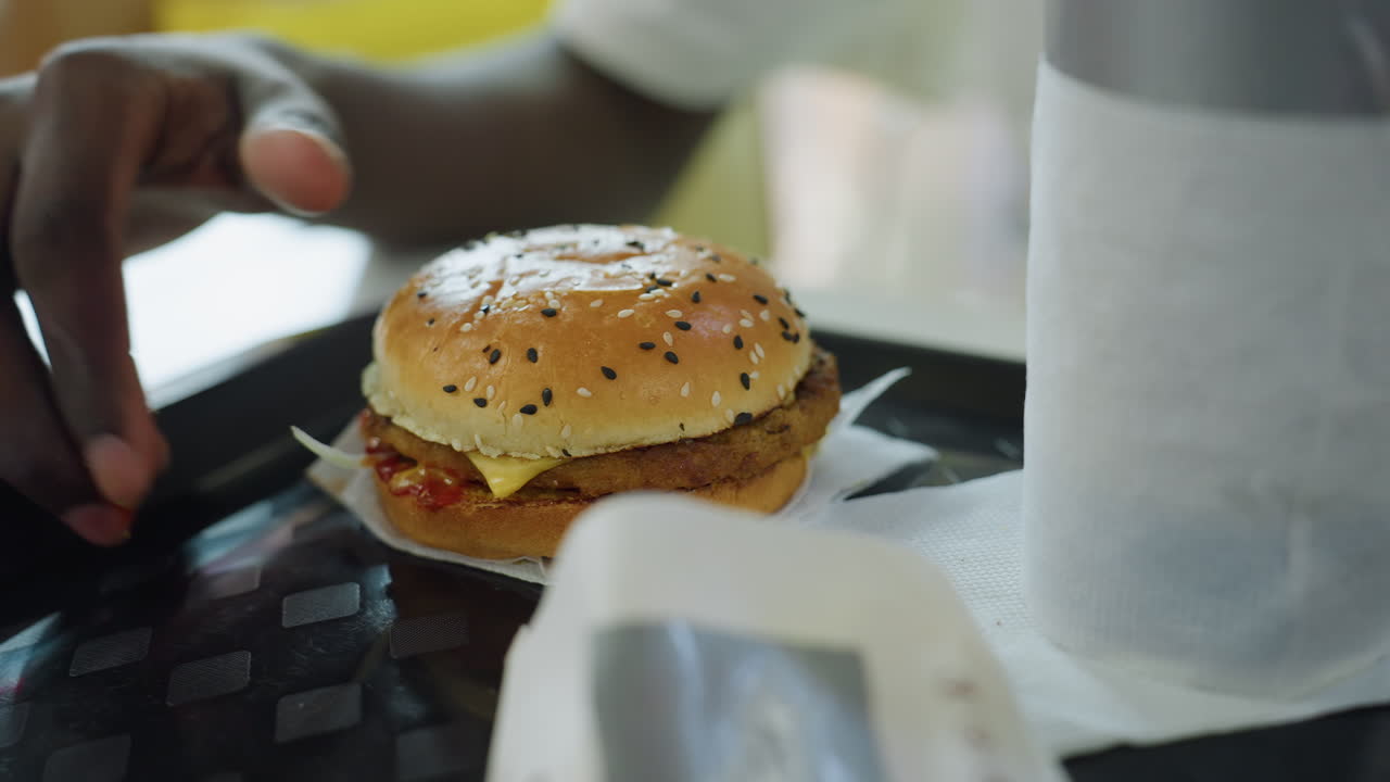 Close up of hand reaching for sesame burger on tray with napkin and drink cup beside fast food packaging indoors under soft light with reflections showing detail of meal preparation and setting moment