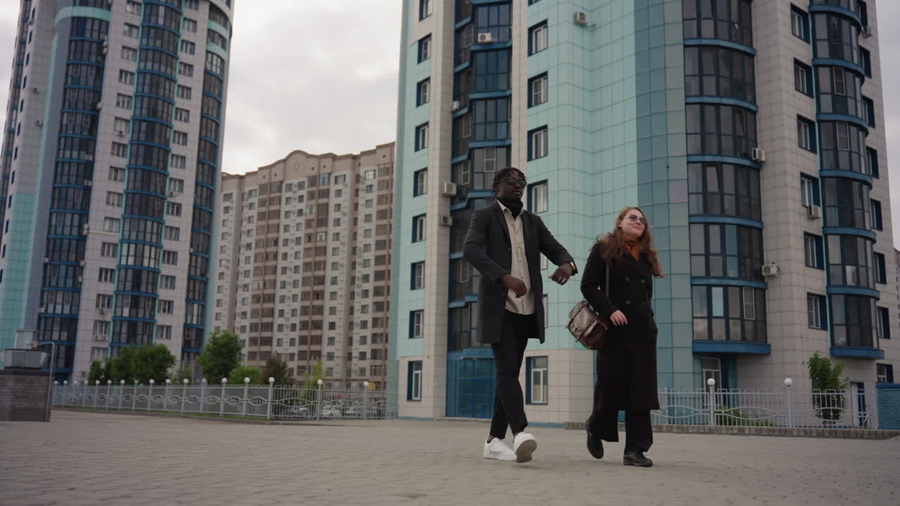 Stylish professionals adjust eyeglasses while walking on stone promenade between tall city towers under cloudy daylight, expressing fashion, and urban elegance through natural synchronized motion