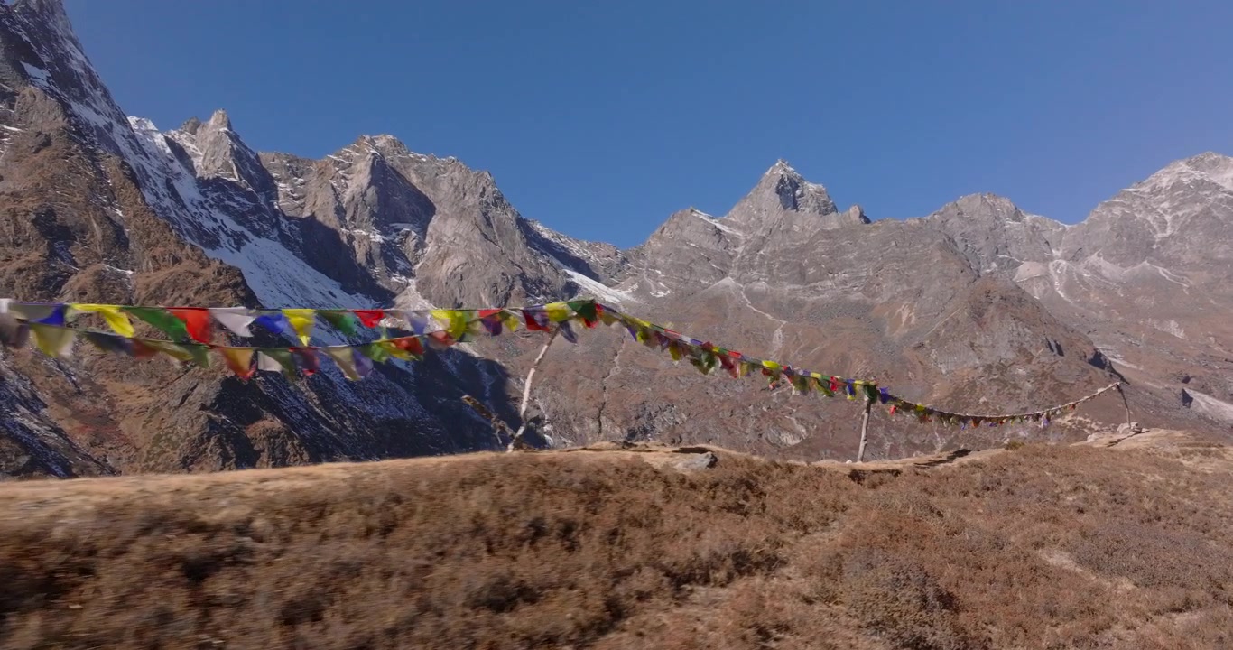 Everest-regionen, Nepal: Svævende luftoptagelse af farverige buddhistiske bedeflag, der blafrer på en bjergryg med Mount Ama Dablam og sneklædte Himalaya-toppe i baggrunden.