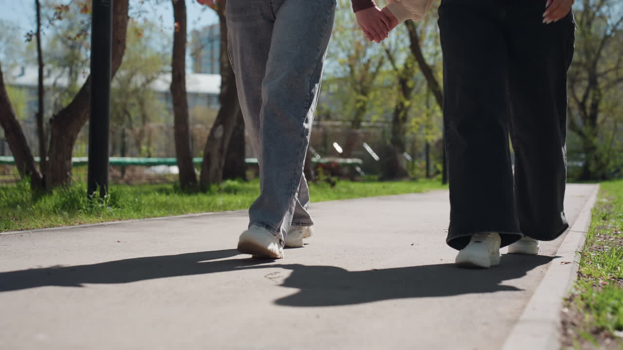 낮은 앵글에서 촬영된 백인 및 아시아 여성들이 청바지와 운동화를 신고 햇볕이 내리쬐는 공원로의 인도에서 발걸음을 맞추어 걷고 있으며, 긴 그림자가 드리워져 있고 나무 줄기가 장면을 둘러싸고 있어요.