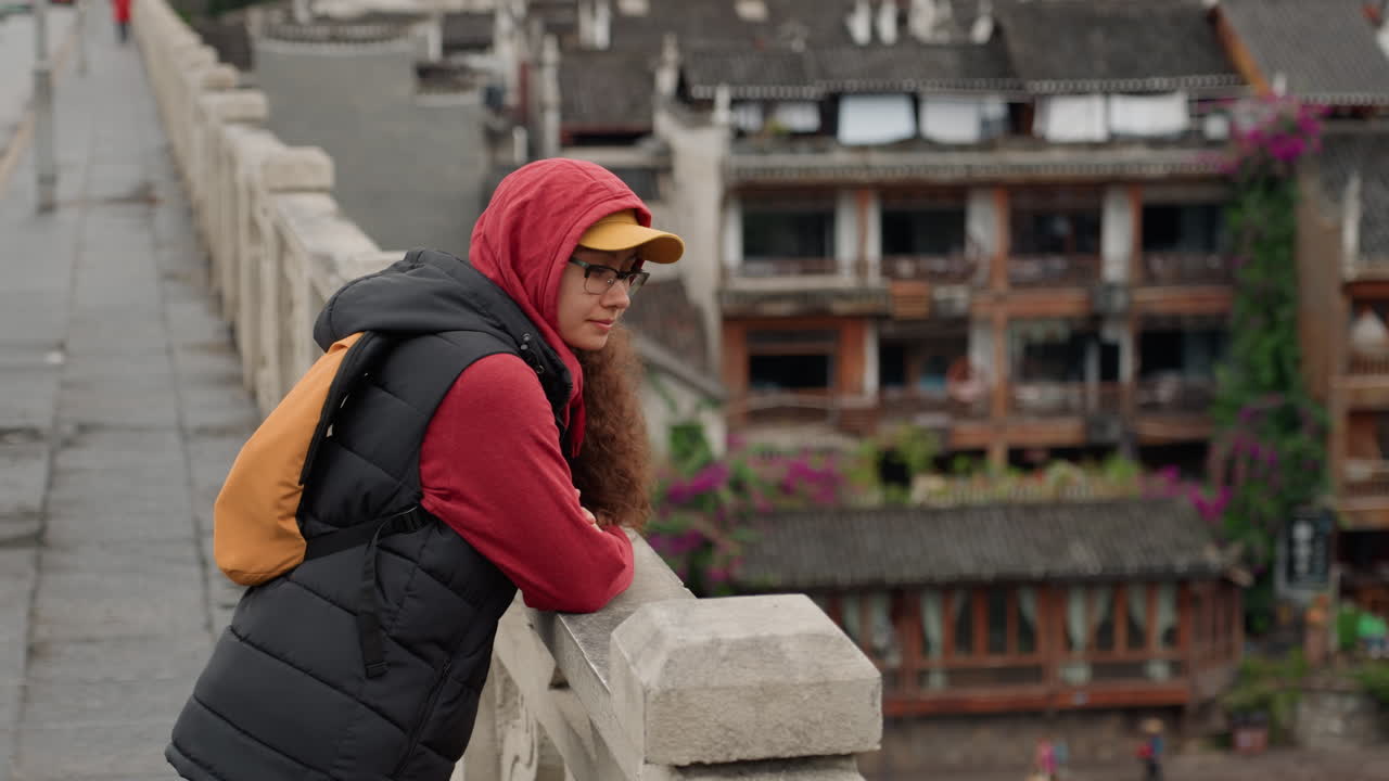Mulher asiática encostada no corrimão da ponte com vista para uma vila ribeirinha, vestindo moletom vermelho, colete acolchoado preto e boné amarelo com mochila laranja, postura contemplativa, parapeito de pedra, varandas de madeira