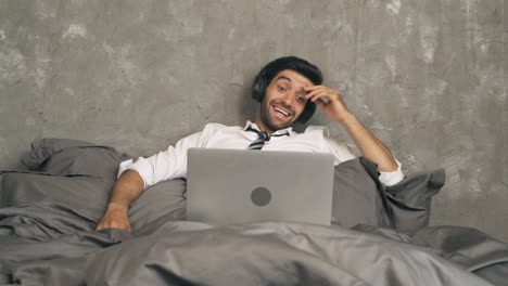 happy business middle east man person watching a funny movie online and laughing on bed in bedroom at home with headphones. activity lifestyle. relax time. computer notebook laptop.