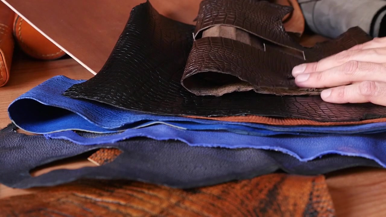 Close up shot of a craftsman examining high-quality genuine leather samples with alligator pattern, blue suede, and brown skin for handmade luxury goods.