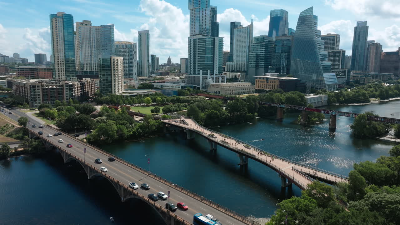 Drone orbits from the south side of Lamar Bridge, capturing both pedestrian and vehicle bridges, then swings east to reveal the full Austin skyline under bright clouds and a clear summer morning sky
