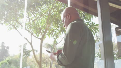 thoughtful african american senior man sitting on balcony in the sun using smartphone