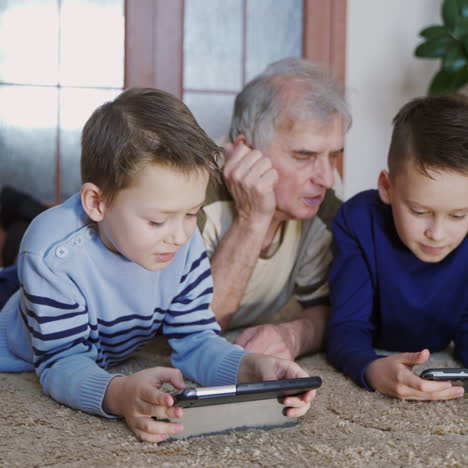 a grandfather with two children are lying and talking on the carpet in the middle of a room in their house. boys have fun with tablets.