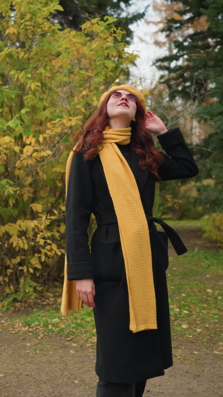 элегантная женщина в черном пальто с желтым беретом и шарфом стоит в осеннем парке, оглядываясь и касаясь ее красных волос с теплой улыбкой