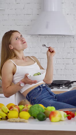 smiling woman eating healthy food. beautiful young lady sitting in the kitchen and eating salad on fresh organic food background. vertical video
