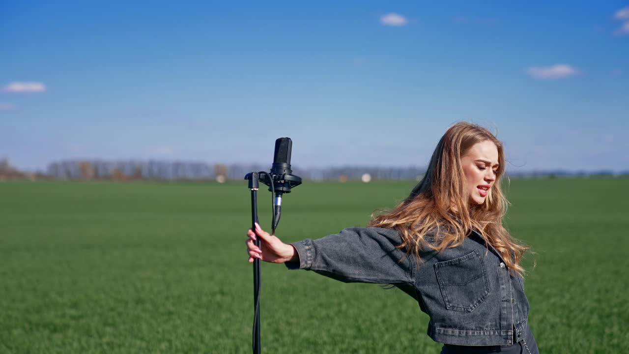 Attractive woman performing on green field background. Beautiful girl in denim suit singing in a microphone and looking at camera outdoors.