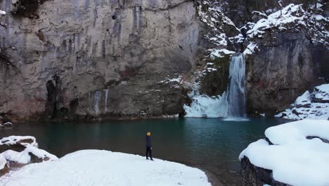 man-walking-towards-waterfall-in-snowy-season,-drone-going-forward-from-the-air-and-approaching-waterfall
