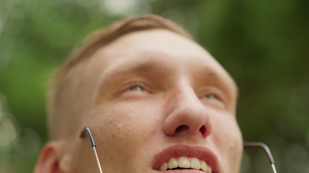 young white man clear glasses upward with hopeful smile, droplets fading, sunlight filtering through leaves, green bokeh background, closeup cinematic perspective, renewed clarity and relief. Frame