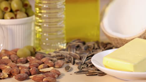 sunflower seeds fall onto a table with almonds, olives, butter, coconut, and oil in bright studio lighting. static camera, vibrant food arrangement