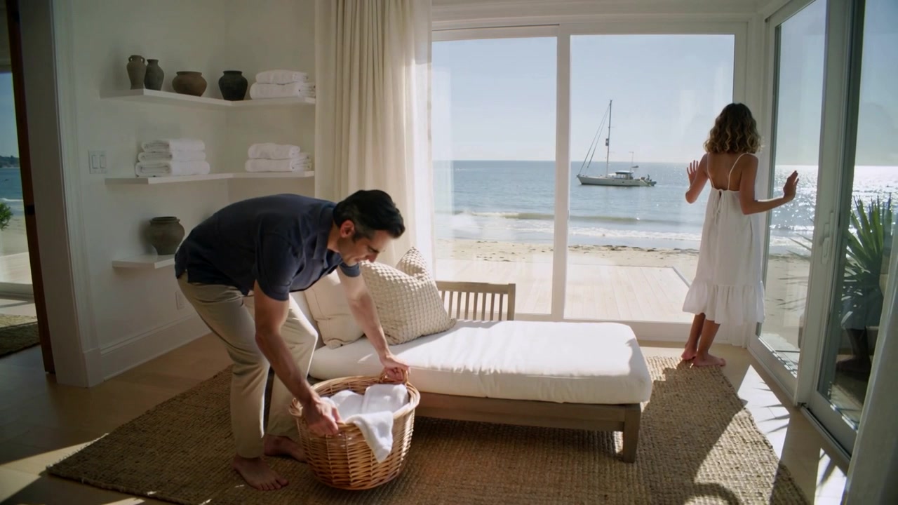 Entering man carrying wicker basket storing linens in sunlit room woman in dress gazing at sailboat