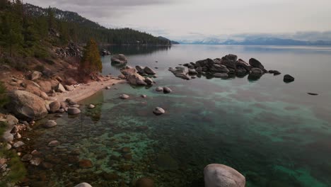 sunny shoreline mid winter day sand harbor secret beach south lake tahoe aerial drone california large boulder rocks crystal clear glassy turquoise fresh water snowy heavenly mountains upwards motion
