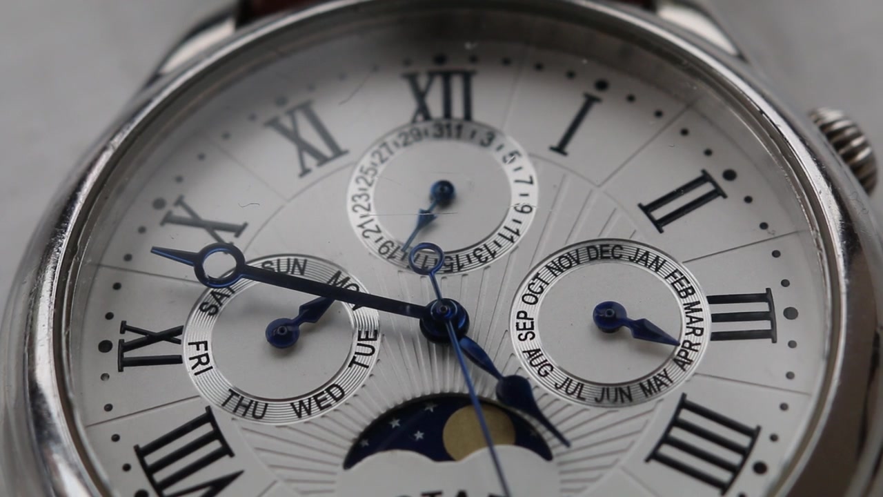 Close up shot showing elegant watch with white dial, blue hands, Roman numerals, calendar subdials and moon phase display in polished metal case