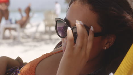 latin girl on beach resort on bright sunny day lowers sunglasses to peer out at view, close up