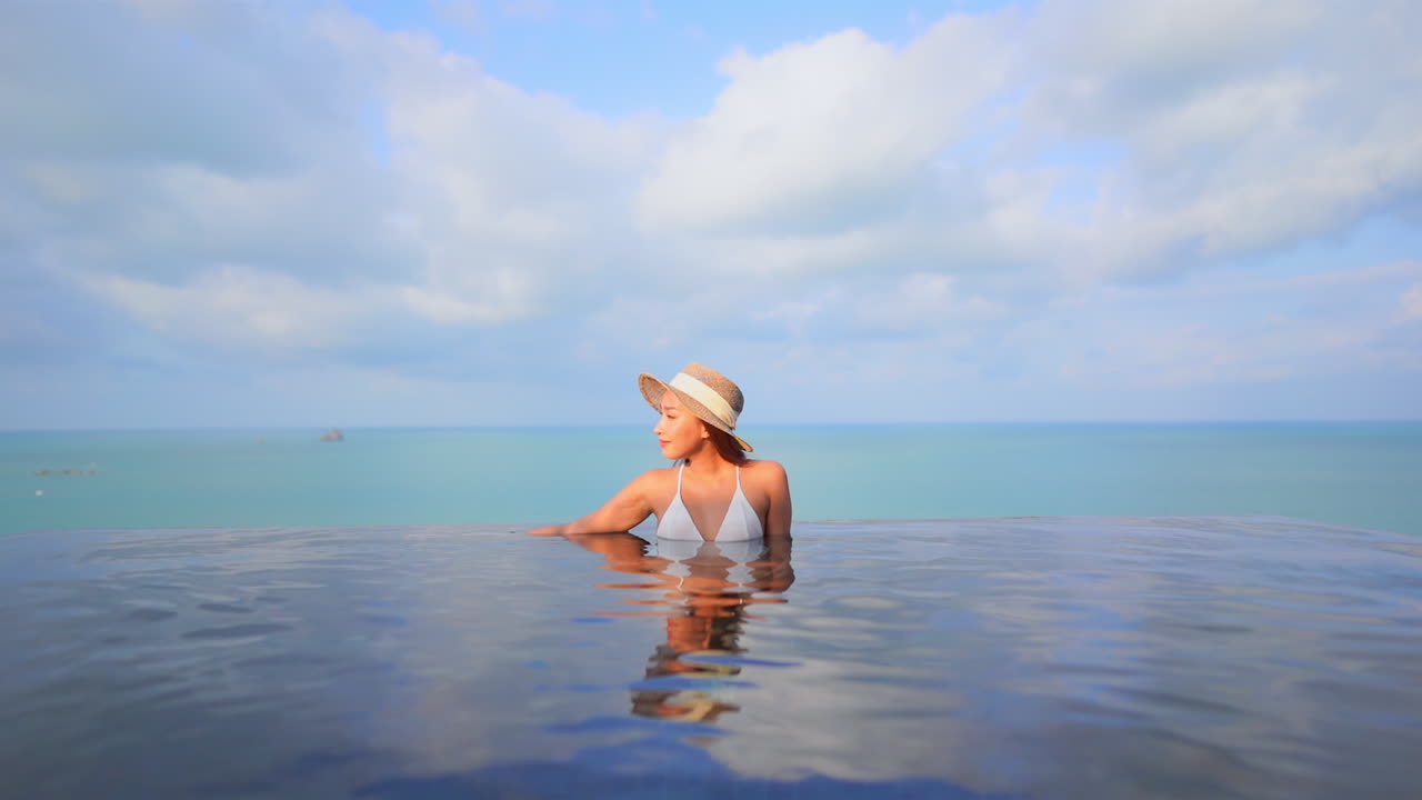 Mit den Ozeanen im Hintergrund entspannt sich eine hübsche junge Frau in brusthohem Wasser am Rande eines Resort-Infinity-Pools.