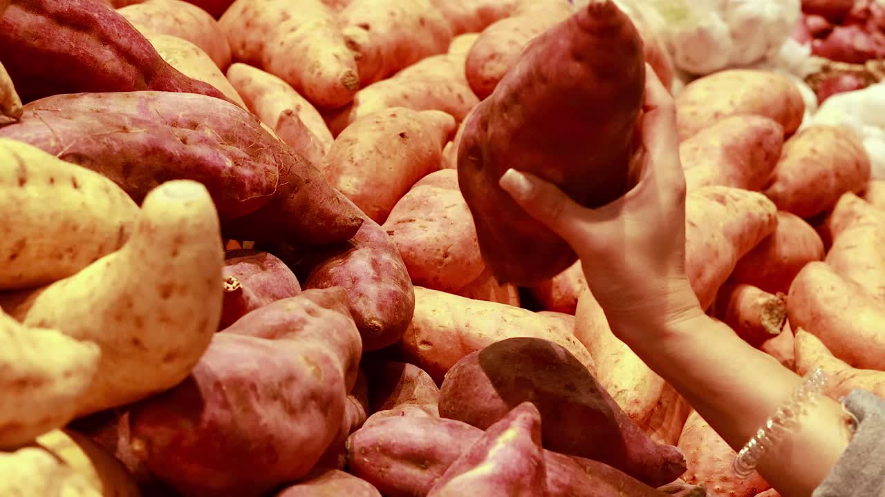 Hands examine sweet potatoes in a well-lit supermarket setting. The focus is on texture and quality selection