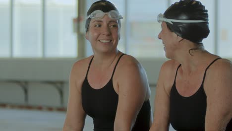 two women in swim gear smiling and talking by a pool