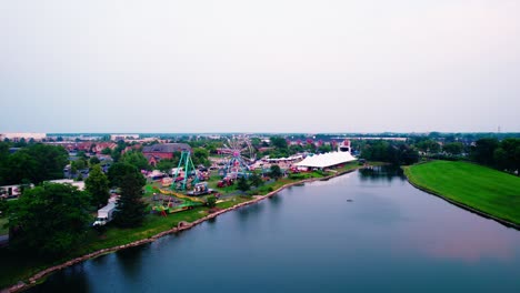 rising-aerial-above-Little-Bear-Lake-towards-Carnival-in-Vernon-Hills,-Illinois,-USA-Century-Arboretum-Park---Ferris-Wheel--Carousell-and-tents,-Aerial-Sunset