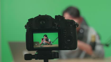 close up de un monitor de cámara grabando hombre asiático con auriculares y computadora revisando teléfono móvil en fondo de pantalla verde