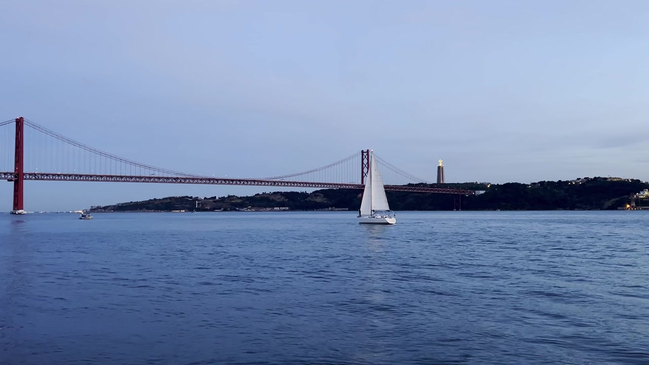 View of the Tagus River with the 25th of April Bridge in the background and a sailboat sailing at dusk. Lisbon. Portugal.