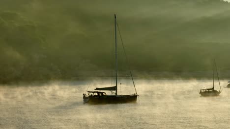 sea fog floats on open ocean surrounding sailboat anchored in open seas at sunrise