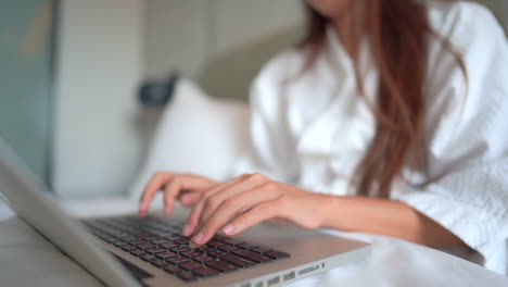 Manos-De-Mujer-Escribiendo-En-El-Teclado-De-Una-Computadora-Portátil-Mientras-Se-Sienta-En-Una-Cama-Con-Albornoz-De-Hotel,-Sin-Rostro