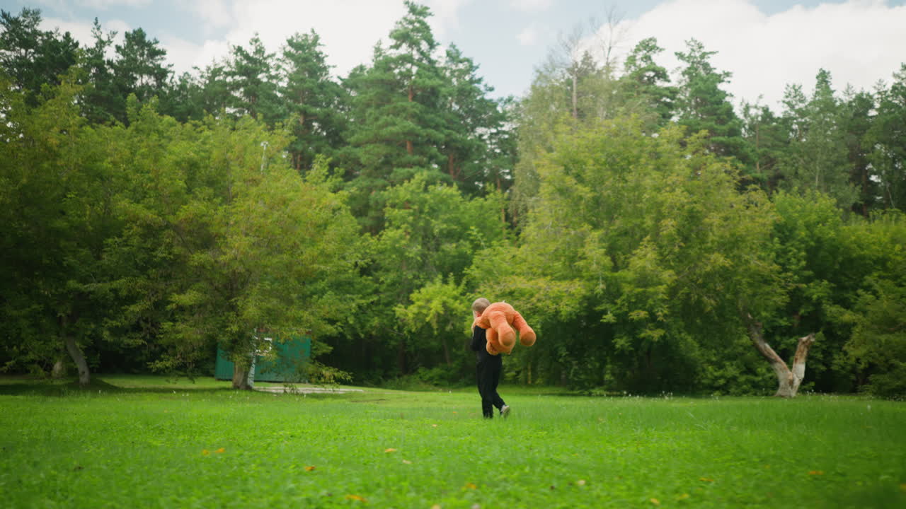 Rear view of young boy holding large teddy bear behind his back while walking across lush green park surrounded by dense trees, evoking playful childhood