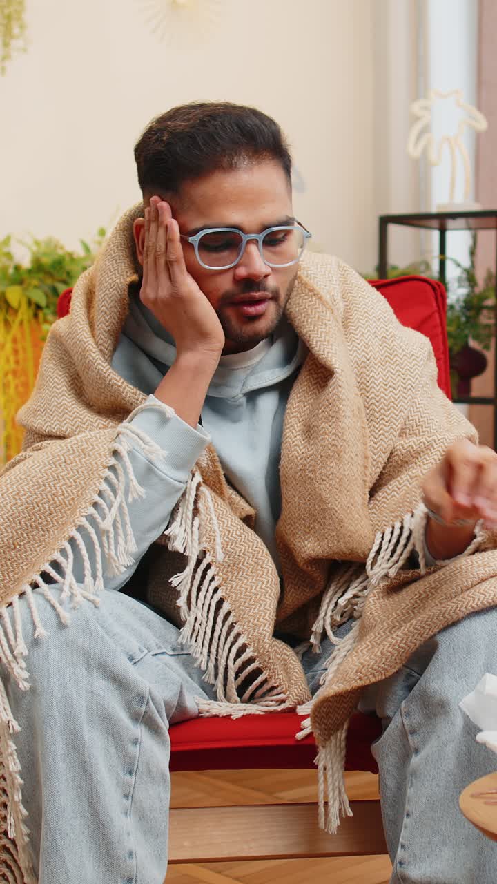 Sick ill indian man suffering from cold or allergy sits on home chair sneezes wipes snot into napkin