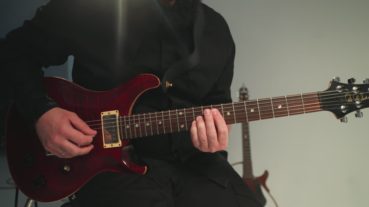 검은 옷을 입은 기타리스트가 회색 바탕에 은 전기 기타를 연주하고 앉아 있으며, 다른 기타가 배경에 보입니다.