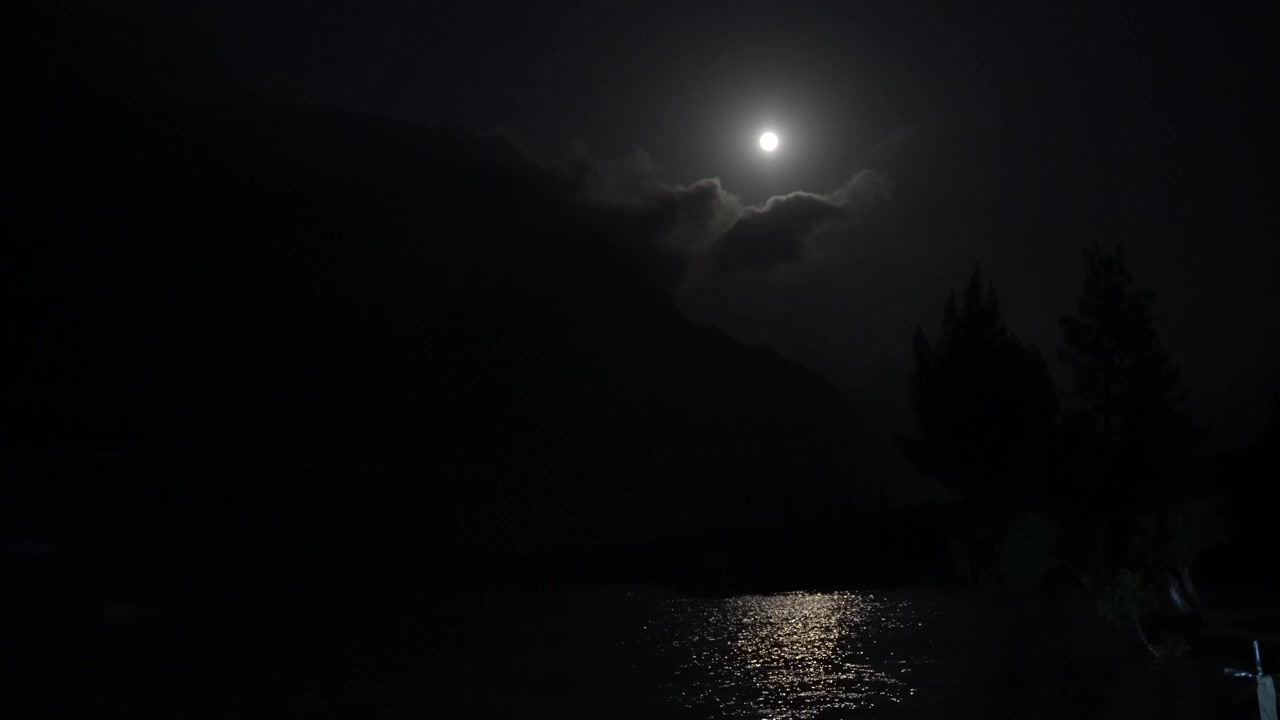 Klar måne skinner gjennom skyer, reflekteres over mørkt vann ved siden av silhuetter av trær om natten