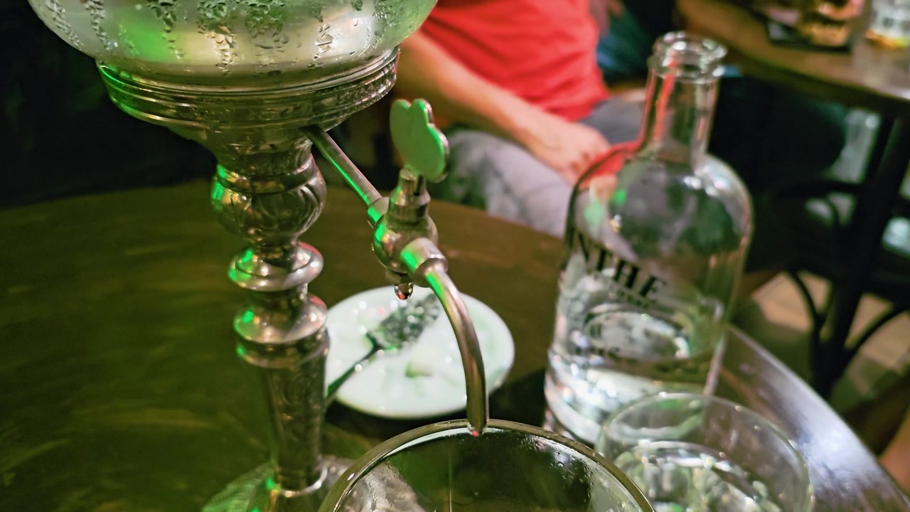 Nahaufnahme des Absinth-Louche, der aus einem Vintage-Brunnenauslauf in ein Glas tropft, mit Spirituosenflasche, Wasserglas, Absinthlöffel, rundem Bartisch und stimmungsvoller grüner Lounge-Beleuchtung.