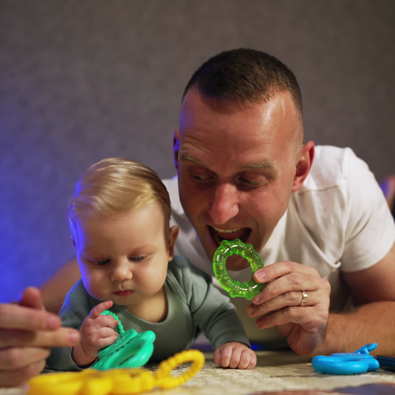 Un papà caucasico e il suo neonato sono sdraiati sul pavimento. Il padre dà un giocattolo per la dentizione al suo bambino