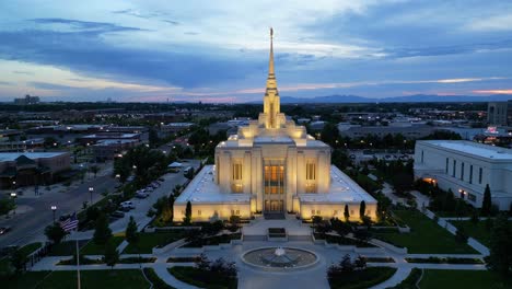 LDS-Mormon-Temple-in-Ogden-Utah-drone-flight-flying-at-dusk-on-beautiful-summer-night-static-symmetric-shot-as-water-fountain-and-American-flag-blowing-in-breeze-and-cars-driving-by-in-the-city