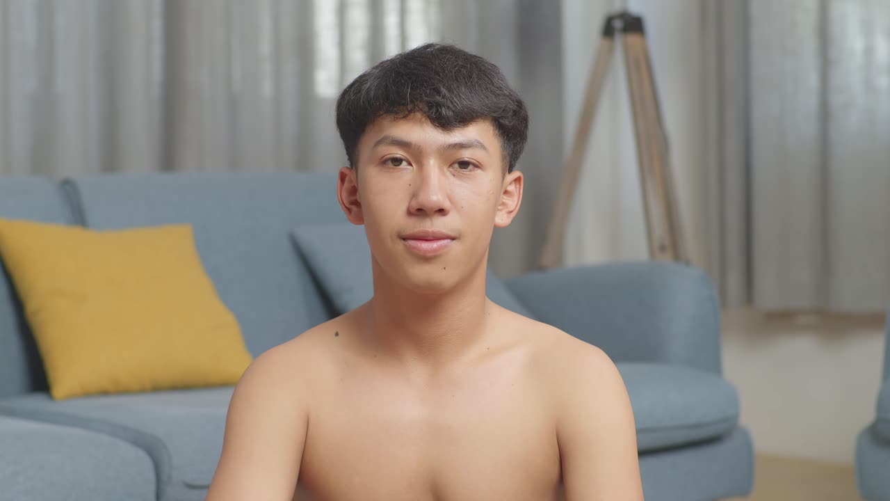 집에서 셔츠를 벗고 운동하는 동안 매트에 앉아 카메라를 향해 미소 짓는 덤벨을 가진 아시아 십대 소년의 클로즈업
