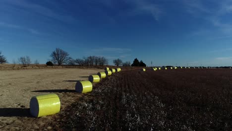 sweeping drone aerial shot of a midwestern cotton farm with fresh bales of harvested cotton wrapped in bright yellow material against a blue open sky