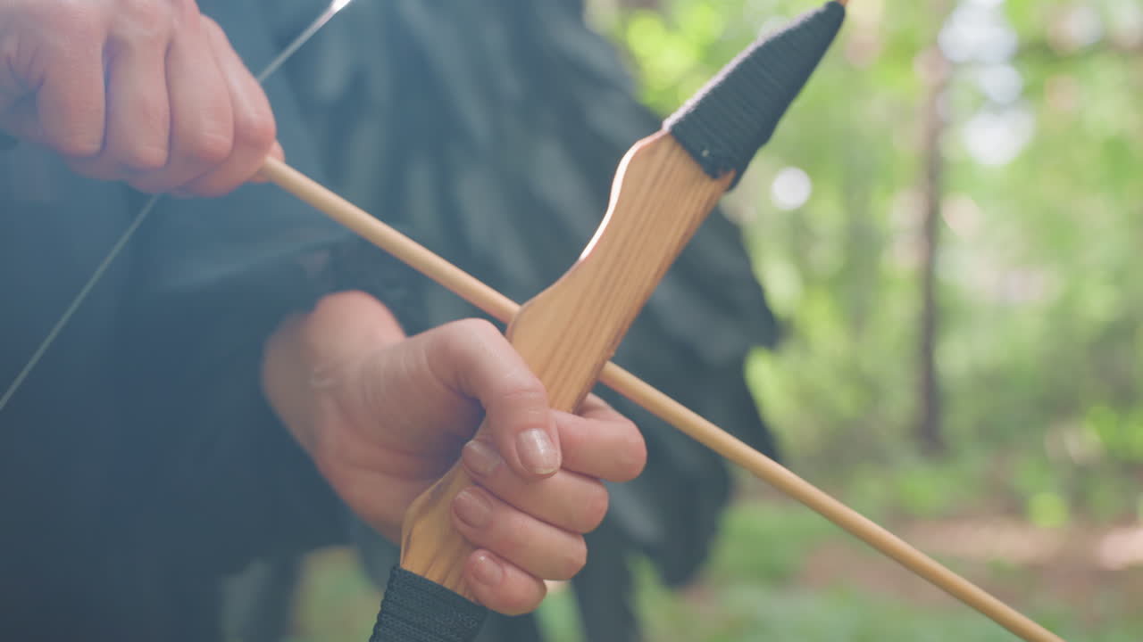 Close up of hands gripping wooden bow with arrow ready to shoot, soft sunlight illuminating fingers and texture, symbolizing focus, readiness, and calm power within green forest atmosphere