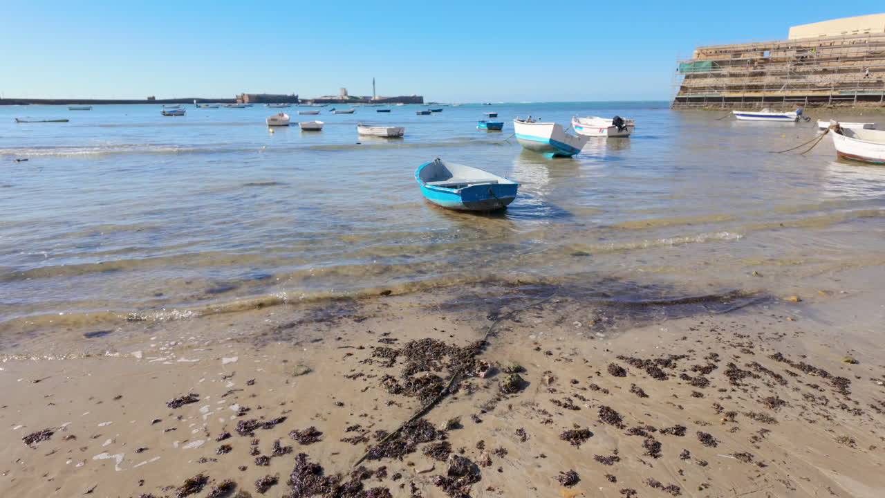 Shallow waters edge on a sandy beach in Cádiz with several small boats floating near the shore, clear blue sky above, and a distant view of the city's maritime structures