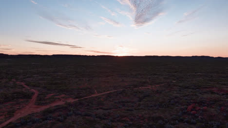 Track-back,-Tilt-up,-Sunset-over-the-outback-hills-of-Mount-Isa-with-a-fork-in-the-red-soil-dirt-road,-blue-and-orange-sky-with-feathery-clouds-and-lens-flares