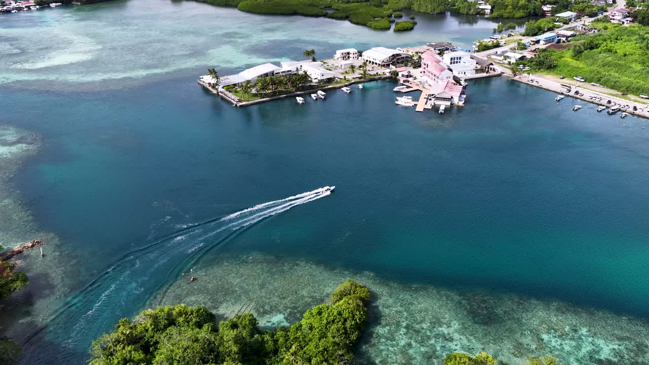 Droneoptagelse i høj vinkel følger en hvid speedbåd, der sejler gennem klart tropisk vand mod en marina.