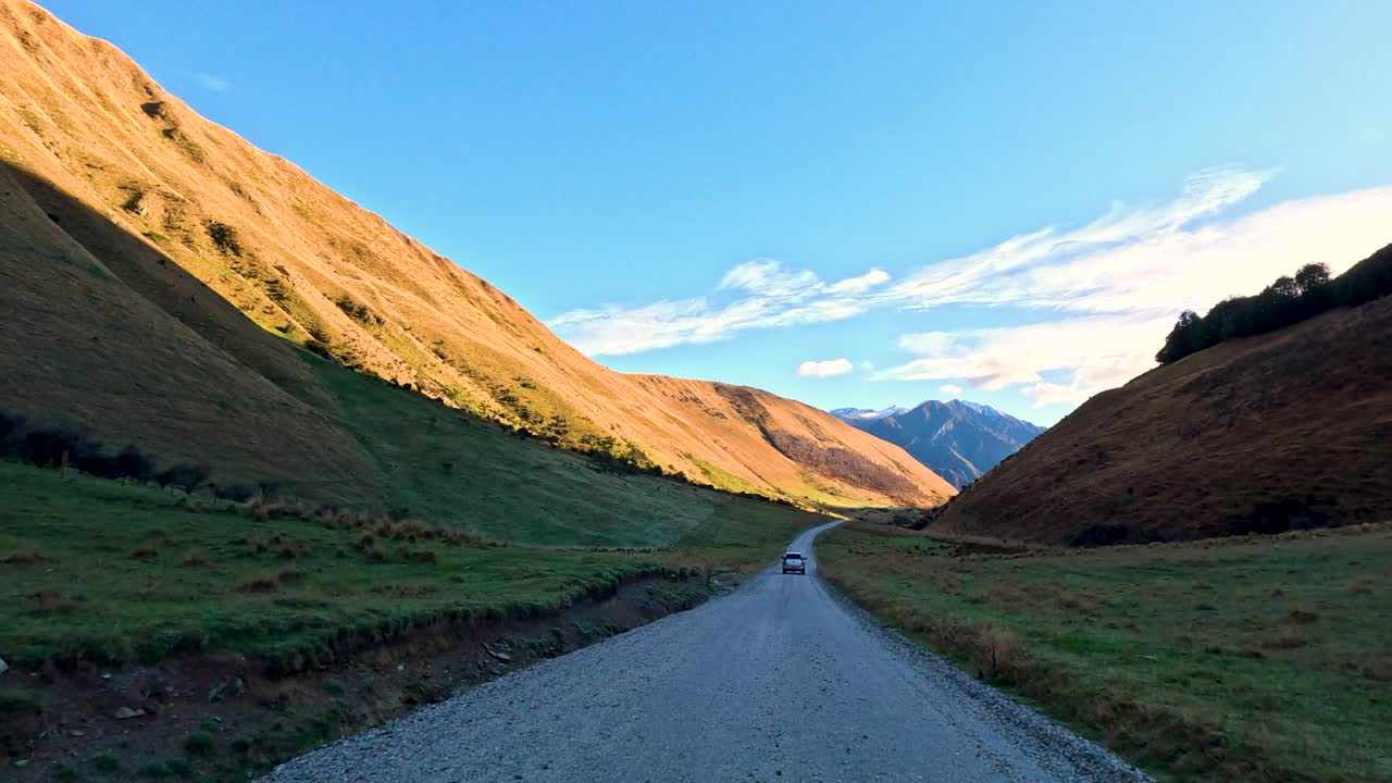 Vehicle travels along gravel road between sunlit hills, wide landscape, steady camera, natural daylight
