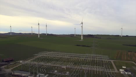 Toma-Aérea-Hacia-Atrás-De-La-Producción-De-Energía-Crítica:-Molinos-De-Viento-Giratorios-En-Un-Campo-Con-Una-Subestación-Eléctrica-En-Primer-Plano-En-Sauerland,-Alemania