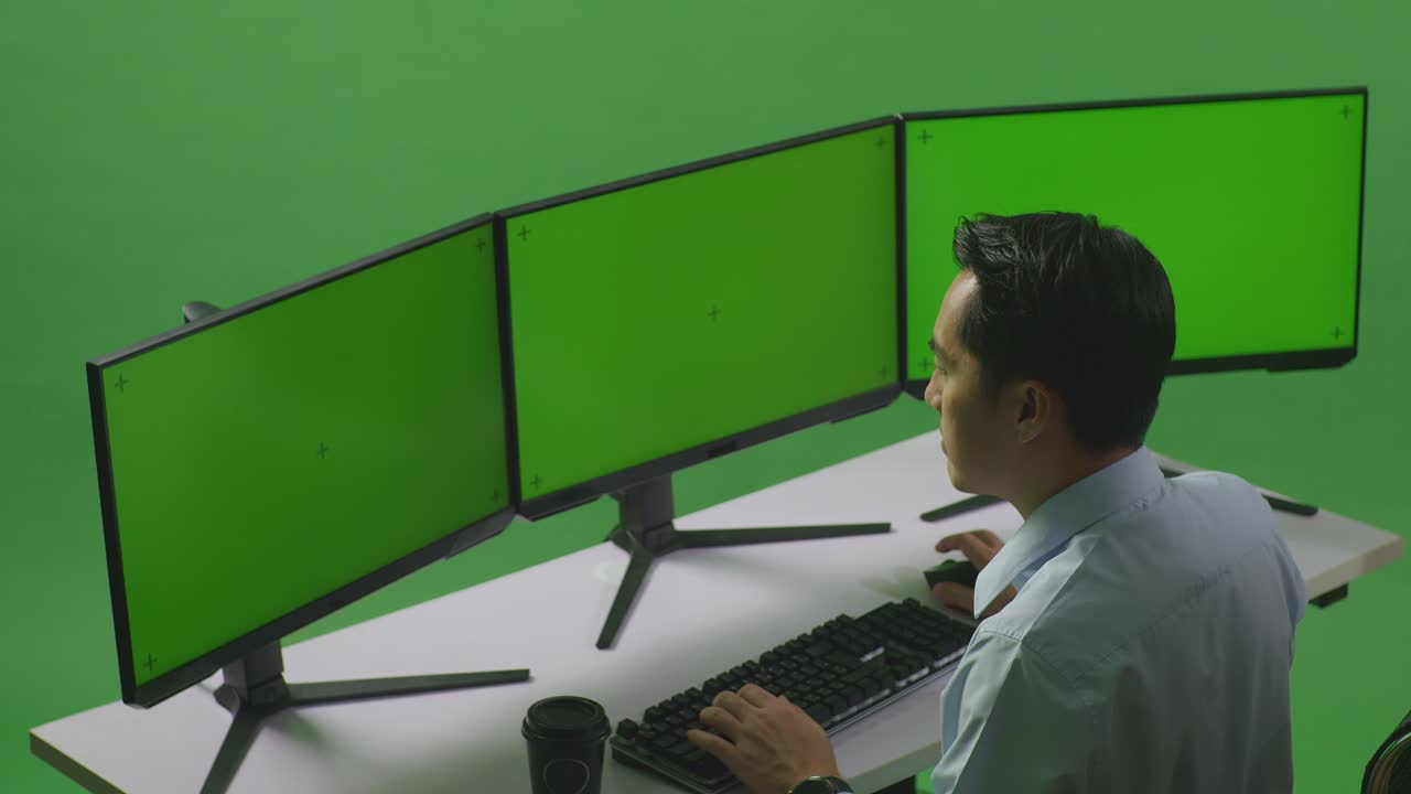 vista lateral de un hombre asiático pensando en algo mientras trabaja con un simulacro de varios monitores de computadora en un estudio de pantalla verde