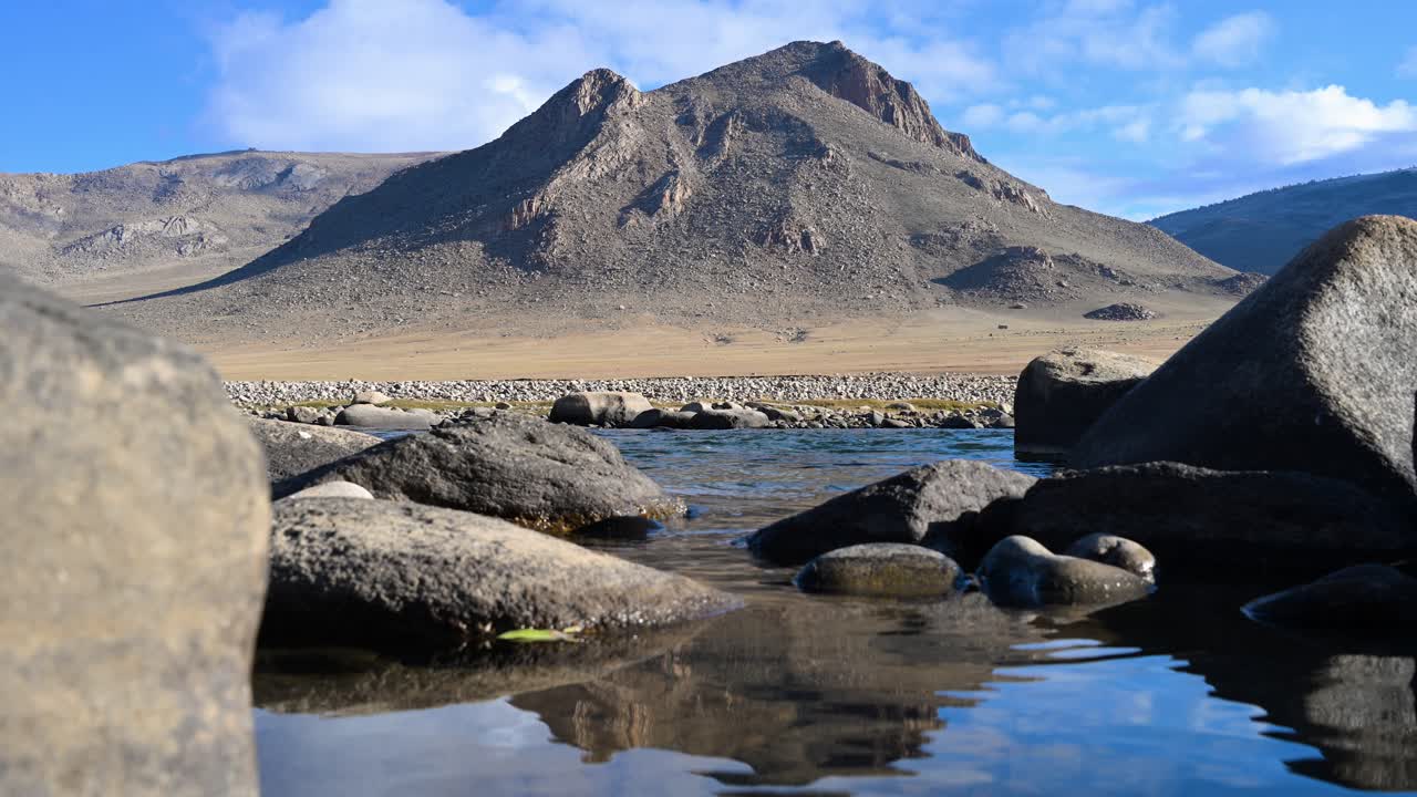 A clear river flows through a remote Mongolian valley, with a majestic, rugged mountain reflected on the water's surface. A serene and scenic autumn landscape