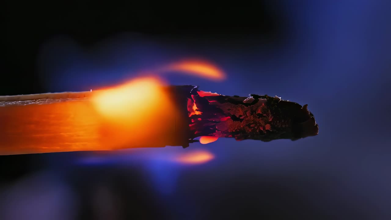 Wooden stick gradually burning, transforming into orange and blue flames while releasing smoke against deep dark background through progressive combustion process