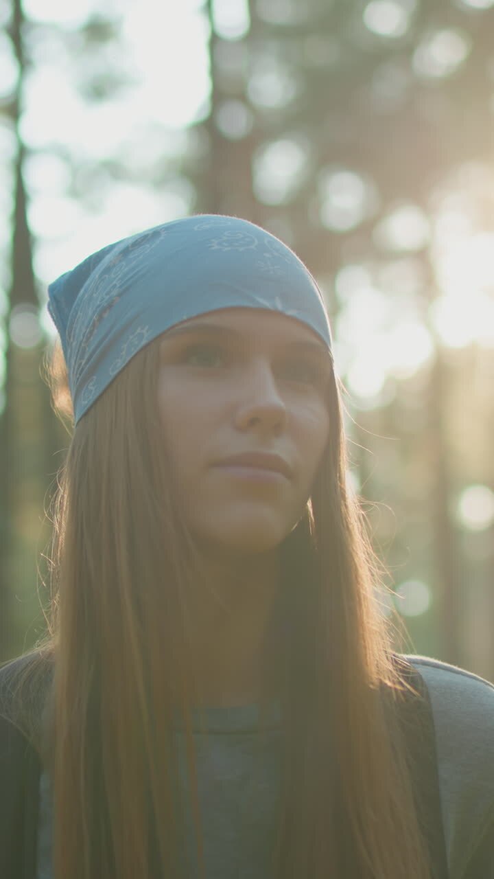 Young woman wearing blue headscarf walking through peaceful forest, looking contemplative with soft sunlight creating a warm glow behind her, surrounded by trees and immersed in nature