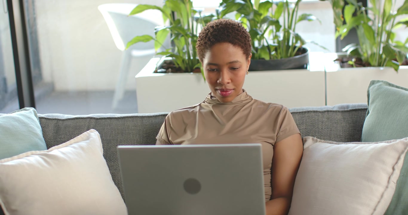 Adult African American woman starting remote work session, sitting on sofa, typing on laptop